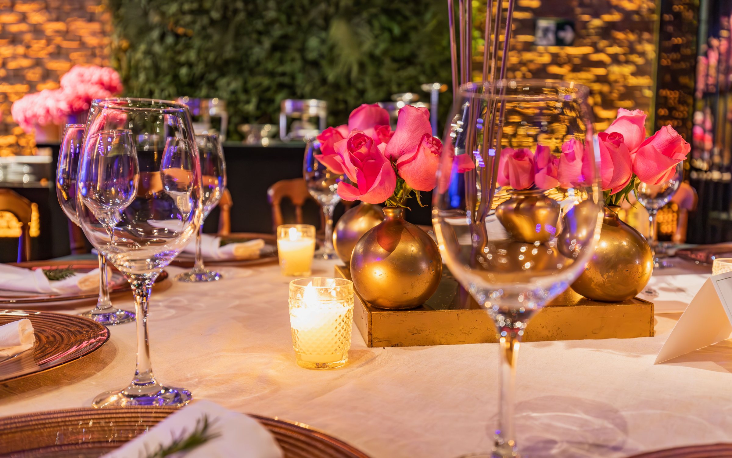 Mesa de evento con copas de cristal, rosas en jarrones dorados y velas encendidas